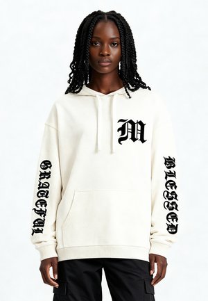 Crème hoodie met een voorste kangoeroezak, voorzien van een groot zwart "M"-logo op de borst en de teksten "GRATEFUL" en "BLESSED" op de mouwen.