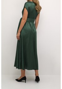 Kvinna står på sidan iförd en ärmlös grön midiklänning med ett brett bälte och svarta högklackade skor mot en enfärgad bakgrund.