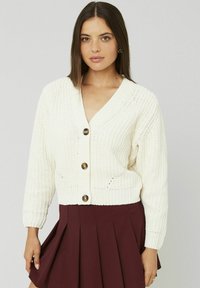 Cardigan en tricot crème avec un décolleté en V profond, présentant des boutons bruns proéminents et une texture côtelée. Associé à une jupe plissée bordeaux.