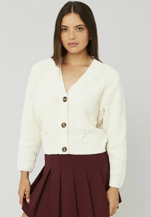Cardigan en tricot crème avec un décolleté en V profond, présentant des boutons bruns proéminents et une texture côtelée. Associé à une jupe plissée bordeaux.