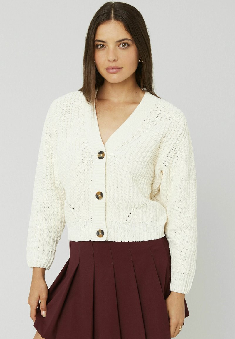 Cardigan en tricot crème avec un décolleté en V profond, présentant des boutons bruns proéminents et une texture côtelée. Associé à une jupe plissée bordeaux.