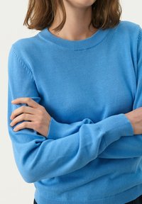 Gaiši zils adīts džemperis ar apaļu kakla izgriezumu, ribotu apročām un gludu tekstūru; pieguloša fasona ar smalku plecu izstrādājumu.