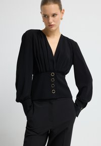 Blusa nera con scollatura a V profonda, spalle plissettate e maniche lunghe. Presenta una vita aderente con bottoni tondi in tono oro. Tessuto dalla texture liscia.