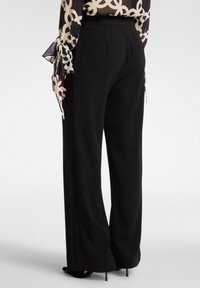 Pantaloni neri a gamba larga con una texture liscia e un elastico strutturato in vita. Sono visibili le tasche laterali; abbinati a una blusa trasparente con motivo.