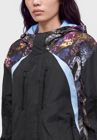 Femme portant une veste noire avec des accents bleus et un imprimé floral et animal multicolore autour des épaules et de la capuche.
