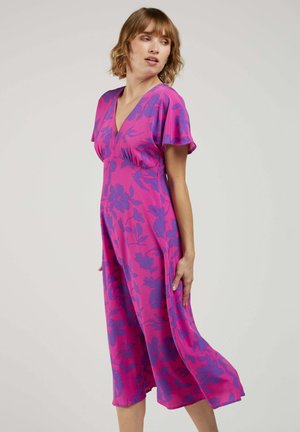 Femme aux cheveux courts portant une robe midi rose avec des motifs floraux violets, debout et regardant sur le côté devant un fond uni.