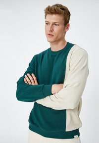 Sudadera color bloqueada en verde y beige con cuello redondo, puños acanalados y tejido ligero. Detalle de la etiqueta en la parte inferior derecha.