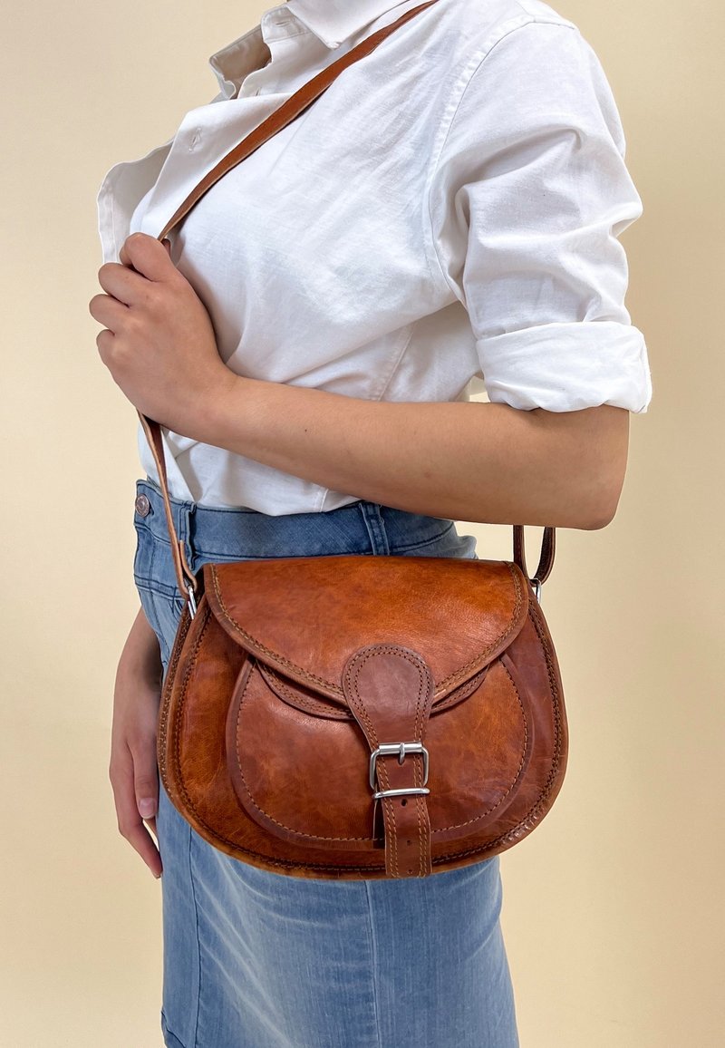 Brązowa skórzana torebka na ramię z zaokrągloną klapką, srebrnym kółkiem jako akcentem i regulowanym paskiem. Gładka faktura z subtelnym połyskiem.