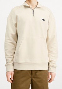 Sudadera beige con cremallera, cuello alto tipo embudo, bolsillo frontal, puños y dobladillo acanalados. Fabricada con un tejido suave y de textura lisa.