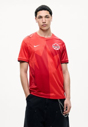 Giovane uomo che indossa una maglia da calcio rossa del Canada con un disegno di foglia di acero e pantaloni neri con una catena, in piedi contro uno sfondo chiaro e semplice.