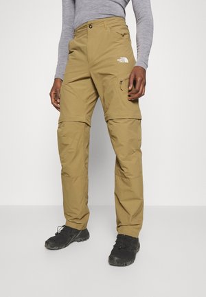 Persoon in tan zip-off cargobroek met het logo van The North Face en zwarte wandelschoenen tegen een witte achtergrond.