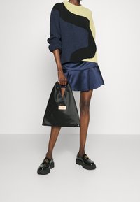Der Pullover hat marineblaue, gelbe und schwarze Strickmuster; kombiniert mit einem blauen, satinähnlichen, ausgestellten Rock; hält eine schwarze Stofftasche mit einem Etikett.