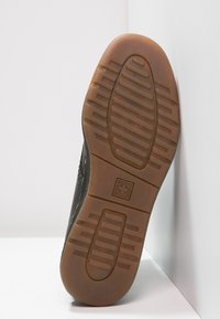 Suela de un zapato negro con suela de goma marrón, que presenta un logo en cruz y patrones de agarre texturizados sobre una superficie blanca.