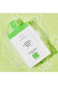 White rectangular bottle with a green cap, labeled "Cocomino Glossing Shampoo." Surrounded by green bubbles on a matching background.