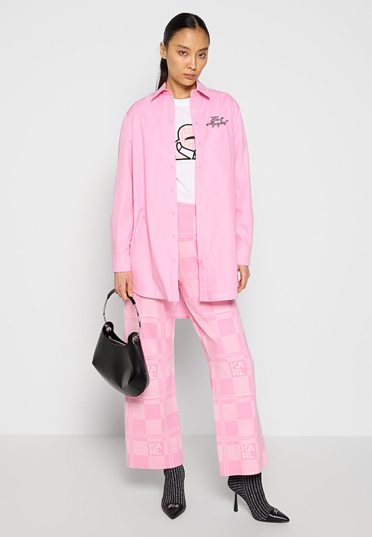 Chemise rose surdimensionnée, T-shirt graphique blanc, pantalon à carreaux roses, sac à main noir et chaussettes noires à motifs avec des talons aiguilles.