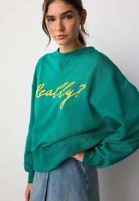 Femeie îmbrăcată într-un hanorac teal cu text galben "Chiar așa?" și fustă din denim deschis albastru, stând cu mâna în buzunar lângă un perete alb.