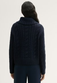 Dunkelblauer Kabelstrickpullover mit hohem Kragen, geripptem Saum und oversized Passform. Strukturiertes Muster auf der Rückseite, weiches Material.