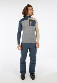 Heren fleece bovenstuk met halve rits in grijs, donkerblauw en beige, met een gestructureerd patroon en een ritszak op de borst. Gepaard met donkerblauwe skibroek.