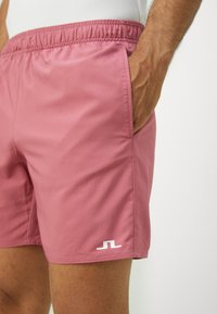Men's pink swim shorts with elastic waistband, side pockets, and white logo detail on the hem. Smooth fabric texture, mid-thigh length.