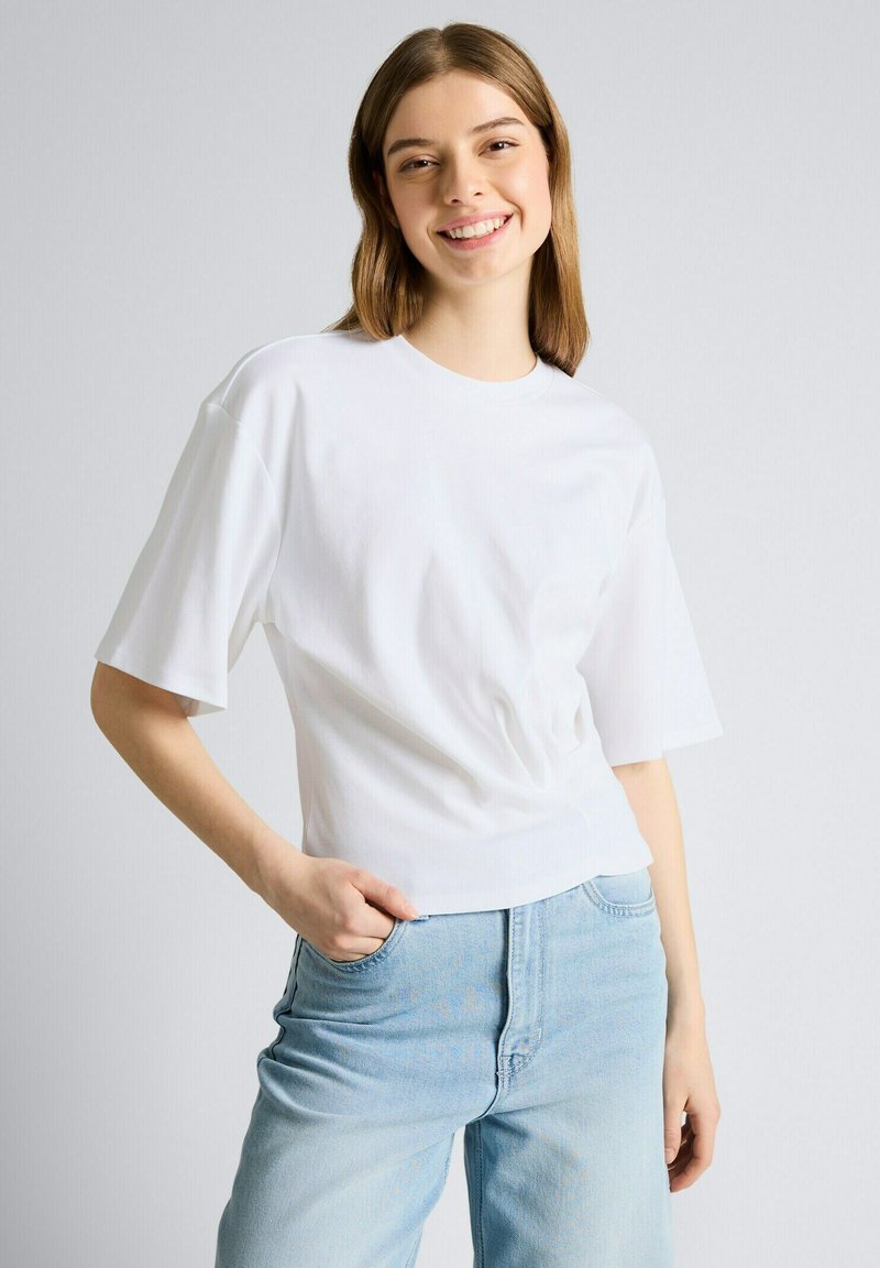 Lächelnde Frau mit hellbraunen Haaren, die ein weißes kurzärmeliges Hemd und hellblaue Jeans trägt, mit einer Hand in der Tasche.