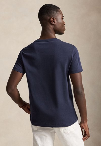 Férfi tengerészkék rövid ujjú ingben és fehér nadrágban, hátulnézetben, semleges háttér előtt.