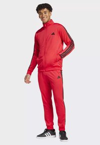 Tuta rossa con strisce laterali nere, giacca con zip e pantaloni abbinati. Presenta il logo Adidas sulla giacca e sui pantaloni, abbinata a sneaker nere.