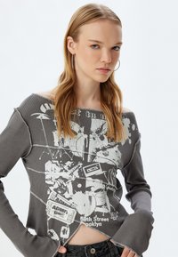 Grau gestreifter Sweatshirt mit Off-Shoulder-Design. Mit weißem Grafikdruck, ausfransenden Kanten und langen, ausgestellten Ärmeln.
