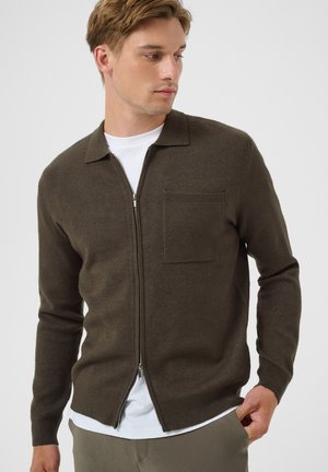 Jeune homme portant un pull zippé marron foncé sur un t-shirt blanc, une main dans la poche, regardant sur le côté.