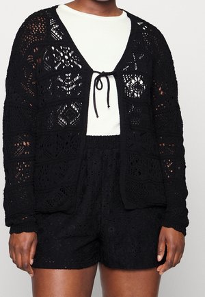 Cardigan long à manches longues en crochet noir, noué devant, porté sur un top blanc, assorti à un short noir en crochet.