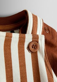 Tuva un brūnas un baltas svītras auduma ar brūnu pogu un daļu no brūnas krekla apkakles zem tās tuvplāns.