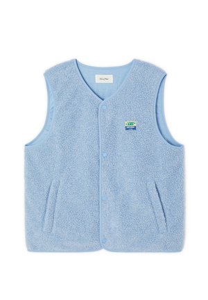 Jeune homme debout portant un gilet polaire bleu clair par-dessus un sweat-shirt gris, un pantalon noir et des baskets blanches sur fond uni.