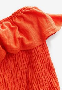 Vêtement en tissu orange vif avec texture élastique froncée et encolure bardot volantée sur fond blanc.