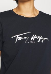 Černé bavlněné triko s metalickým stříbrným logem, na kterém je nápis "Tommy Hilfiger" a "est. 1985" ve stylizovaném písmu.