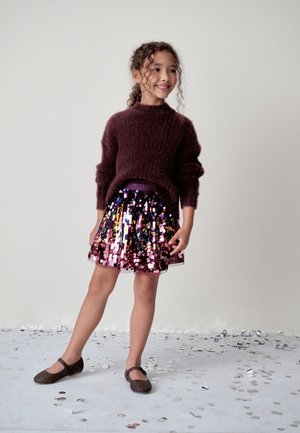 Bordeauxkleurige pluizige trui, iriserende paillettenrok met roze en paarse tinten, en bruine Mary Jane schoenen op een kind dat staat met een confetti-achtergrond.