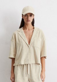 Camicia cropped in terry beige con maniche corte, chiusura a bottoni e taschino sul petto. Abbinata a un cappellino beige coordinato.