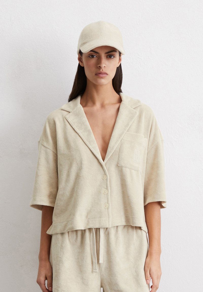 Camicia cropped in terry beige con maniche corte, chiusura a bottoni e taschino sul petto. Abbinata a un cappellino beige coordinato.
