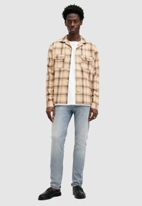 Camisa a cuadros beige con cierre de botones, dos bolsillos en el pecho, usada sobre una camiseta blanca. Jeans de color azul claro y zapatos negros con suela texturizada.