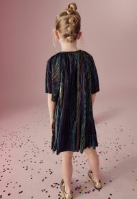 Schimmerndes schwarzes Kleid mit kurzen Ärmeln, mit plissiertem Design, das mehrfarbige metallische Streifen aufweist. Goldene Schuhe und Konfetti auf einer pinken Oberfläche.