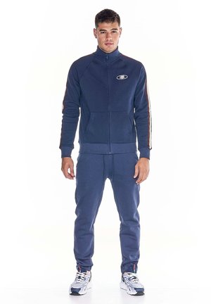 Un homme porte un survêtement bleu marine composé d’une veste zippée, d’un pantalon assorti et de baskets blanches, sur un fond blanc uni.
