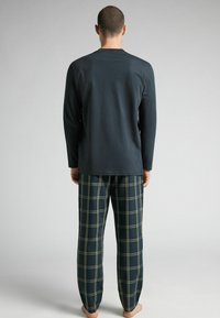 Man som står barfota med ryggen mot kameran, klädd i en mörk långärmad skjorta och marinblå rutiga pyjamasbyxor mot en enfärgad bakgrund.