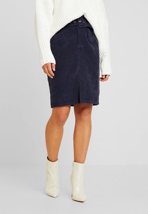 Mujer con un suéter de punto blanco, falda de pana azul marino con cinturón y botas blancas hasta el tobillo, de pie frente a un fondo gris liso.