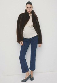 Vrouw draagt een donkerbruine bontjas, beige top, blauwe cropped jeans en zwarte schoenen met lage hak, staand tegen een effen lichte achtergrond.