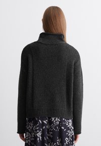 Pull en maille gris foncé avec un col montant, des épaules tombantes et un ourlet côtelé. Le dos présente une finition texturée, porté avec une jupe à motif floral.