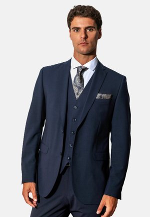 Man met kort krullend haar, gekleed in een donkerblauw driedelig pak, wit overhemd en een stropdas en pochet met paisley-motief.