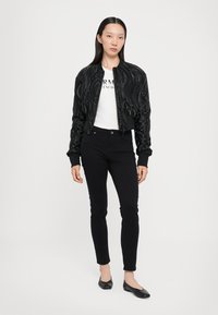 Giacca bomber nera con texture a trapunta, chiusura con zip e polsini a costine, indossata sopra una maglietta bianca e jeans skinny neri. Ballerine nere completano il look.