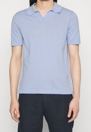 Mann trägt ein hellblaues kurzärmeliges Poloshirt und schwarze Hose, steht vor einem einfarbigen hellen Hintergrund.