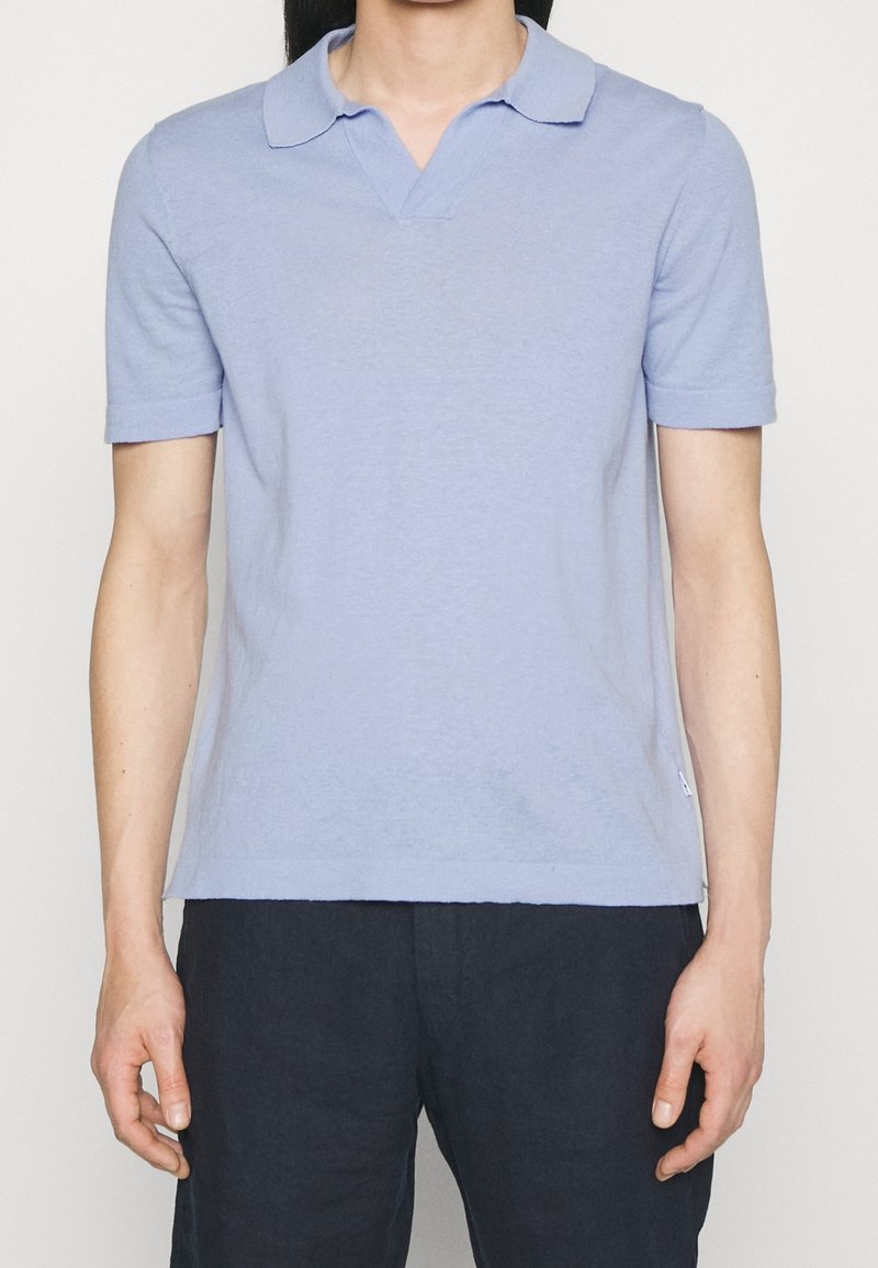 Homme portant un polo bleu clair à manches courtes et un pantalon noir, debout devant un fond clair uni.