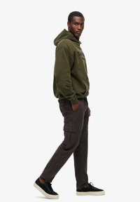 Sweat à capuche vert olive avec logo, pantalon cargo gris foncé avec poches latérales, et baskets noires avec semelles blanches.