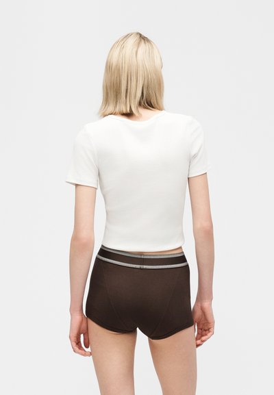Ξανθό άτομο που φοράει ένα λευκό κοντομάνικο τοπ και σκούρα καφέ σορτς με ψηλή μέση, στέκεται δίχως να βλέπει προς το μέρος μας σε απλό φόντο.