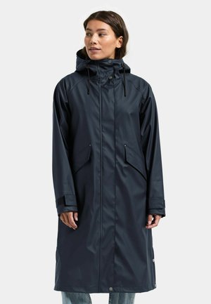 Frau trägt einen langen, dunkelblauen wasserdichten Regenmantel mit Kapuze, Klappentaschen und Handgelenkriemen, steht vor einem schlichten hellen Hintergrund.